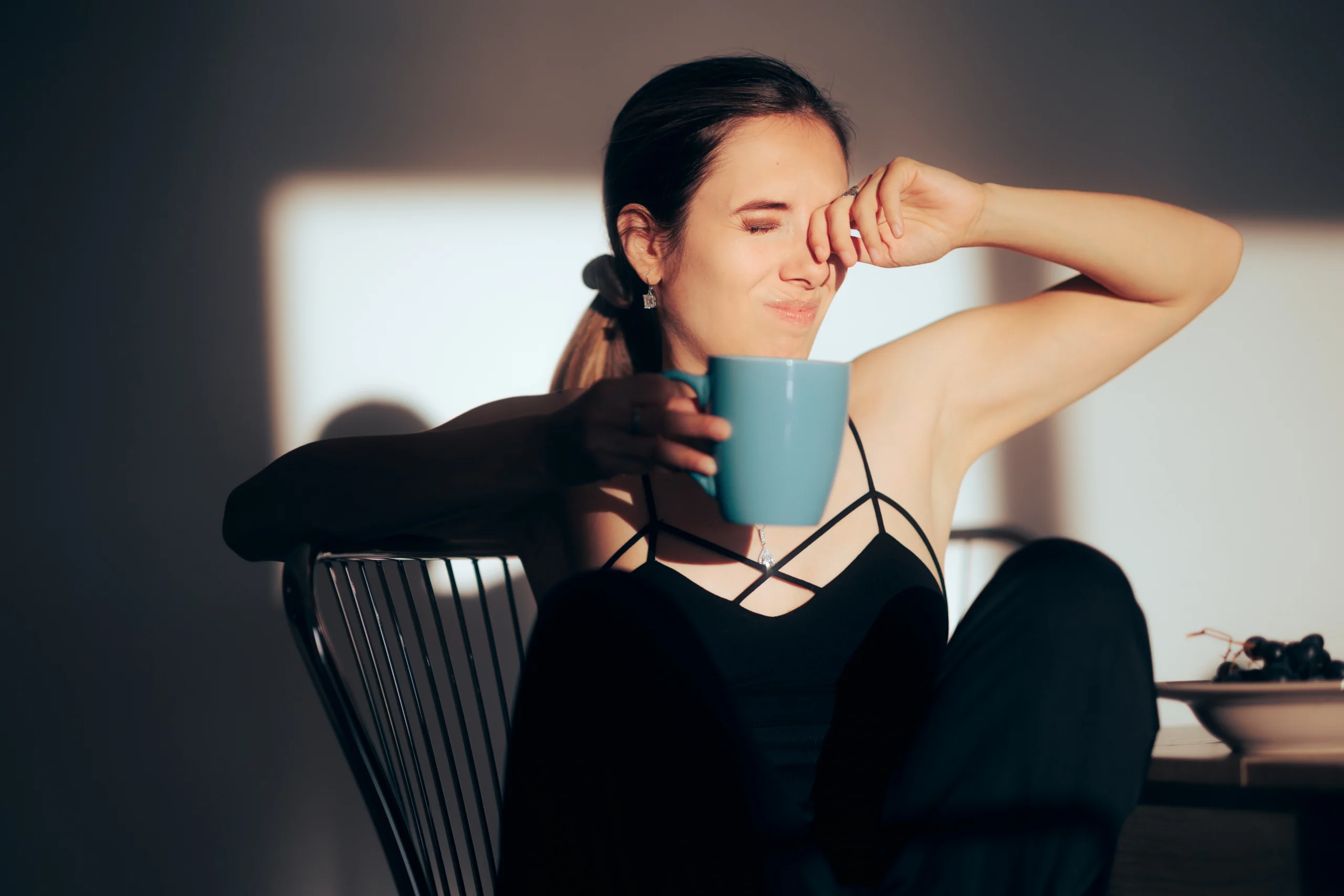 A young woman holding a coffee copy squints and rubs her eyes at her sunny window.