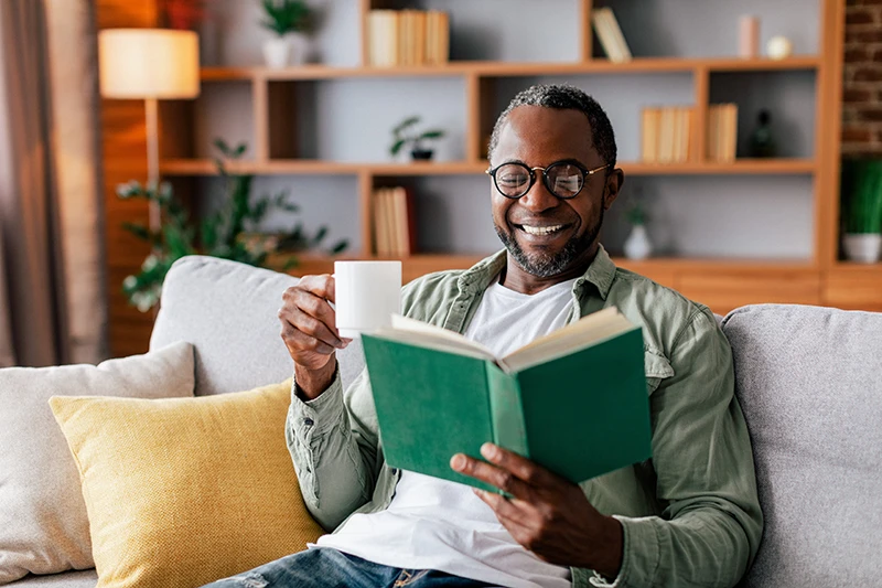 a man reading a book and drinking coffee on a sofa at home